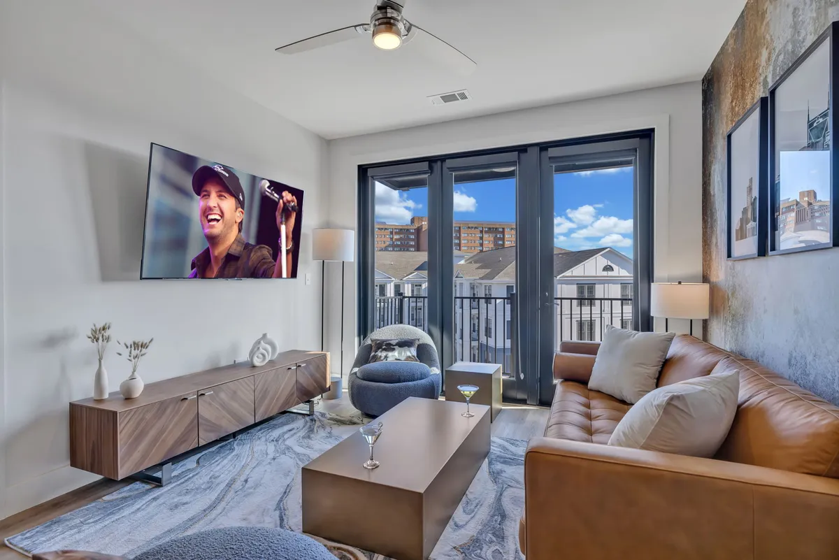 A cheerful, relaxed living area with a comfortable leather sofa, stylish coffee table, and a large television. The space is visually extended by a balcony view through floor-to-ceiling windows, emphasizing the natural light and connection to the cityscape.