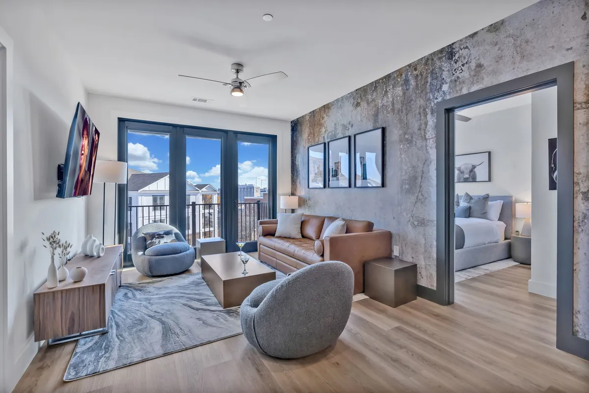 This image shows a contemporary living room with a comfortable sofa, armchair, and coffee table, all arranged to maximize the view of the balcony and the city beyond. The space features modern décor and large windows that allow natural light to flood in, visually connecting the inside with the outdoor balcony space.