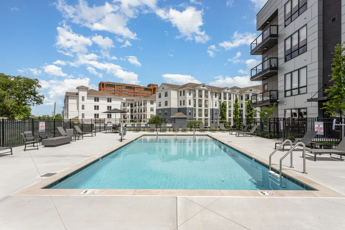 A spacious outdoor pool area featuring sparkling blue waters, lounge chairs, and lush greenery framed by surrounding modern buildings. The relaxed atmosphere suggests opportunities for swimming, sunbathing, and socializing poolside, both during the day and at sunset.