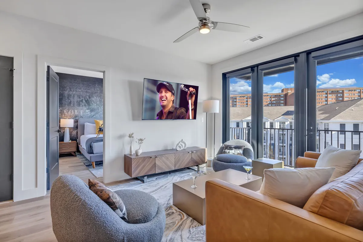 A chic living room with modern furniture, large windows letting in natural light, and a television showing a performer. Stylish decor enhances the atmosphere, and the space feels both lively and inviting.