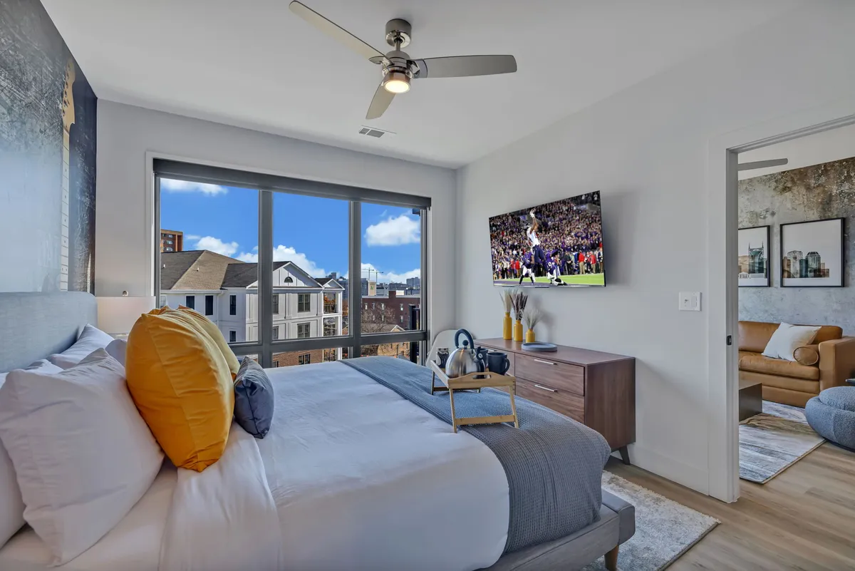 A stylish bedroom with a plush king-size bed, decorative pillows, a large window offering city views, and a TV mounted on the wall. The room feels cozy and modern, with ample natural light and a peaceful retreat vibe.