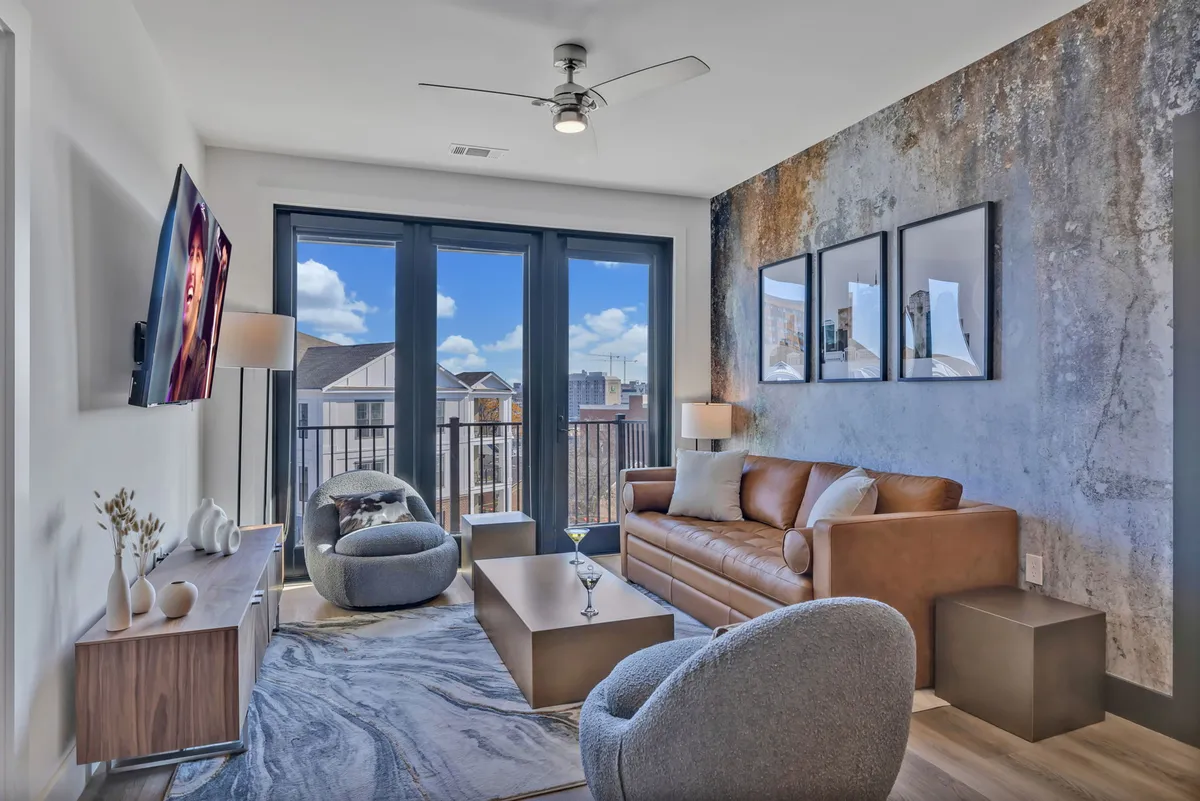 The image showcases a chic, contemporary living room with a plush leather sofa, modern coffee table, bold artistic accents, and expansive windows overlooking the Nashville skyline at sunset. The space radiates warmth and style, inviting guests to relax or entertain with city views as their backdrop.