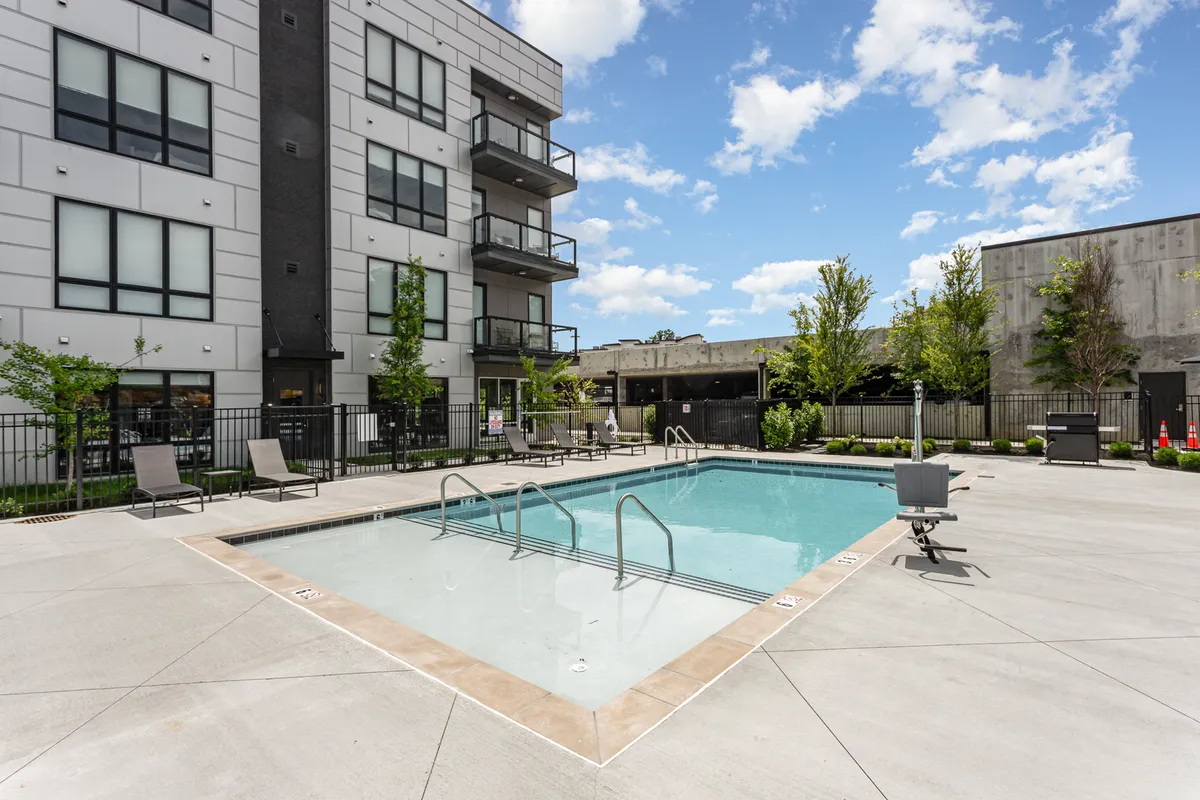 A modern apartment building with balconies overlooks a sparkling swimming pool set in a landscaped courtyard. The scene suggests a relaxed social atmosphere, ideal for evening swims or group gatherings by the water.