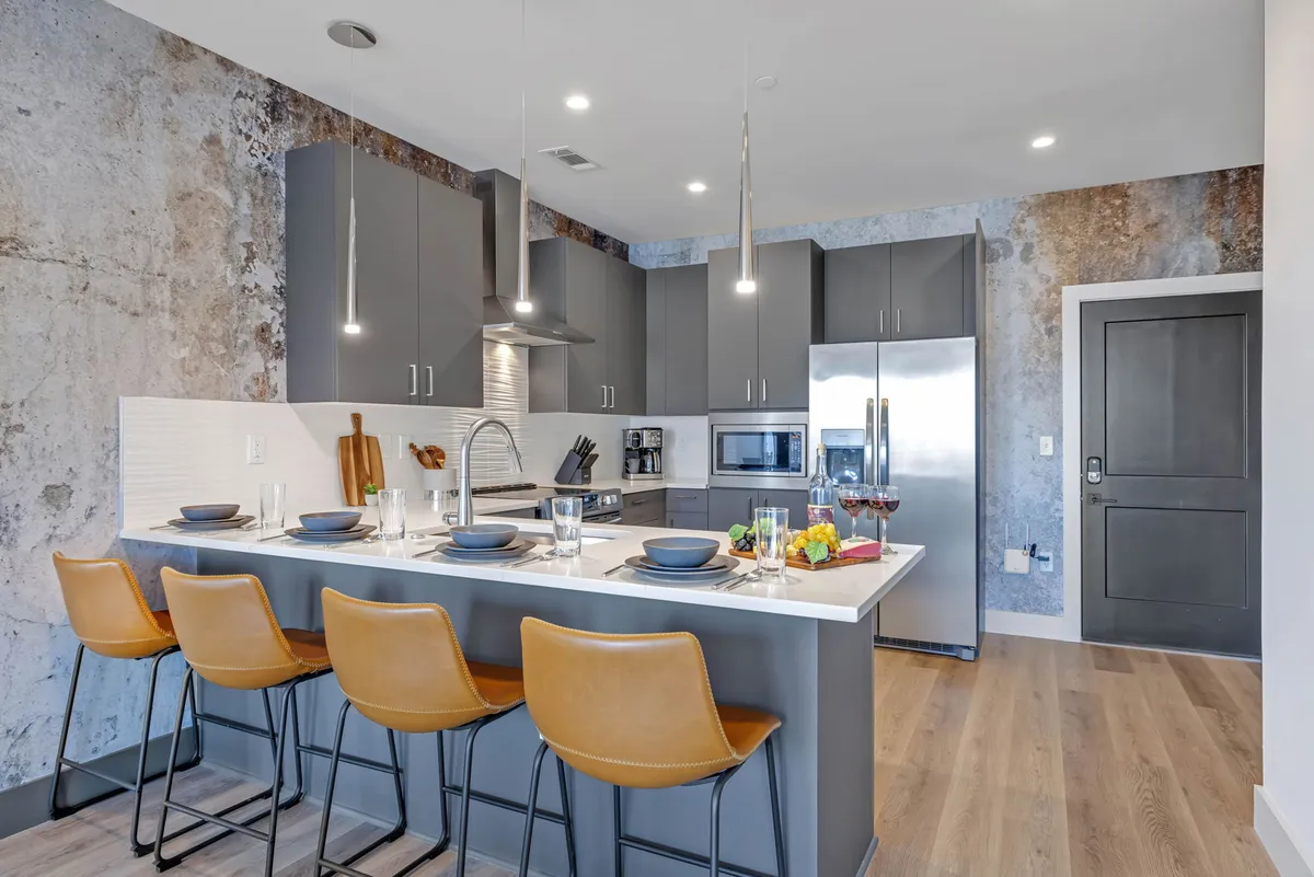 A bright, contemporary kitchen featuring a large island with bar stools, stainless steel appliances, and a textured wall. The kitchen opens up to adjacent living areas, reinforcing the feeling of seamless flow between the cooking, dining, and social spaces.