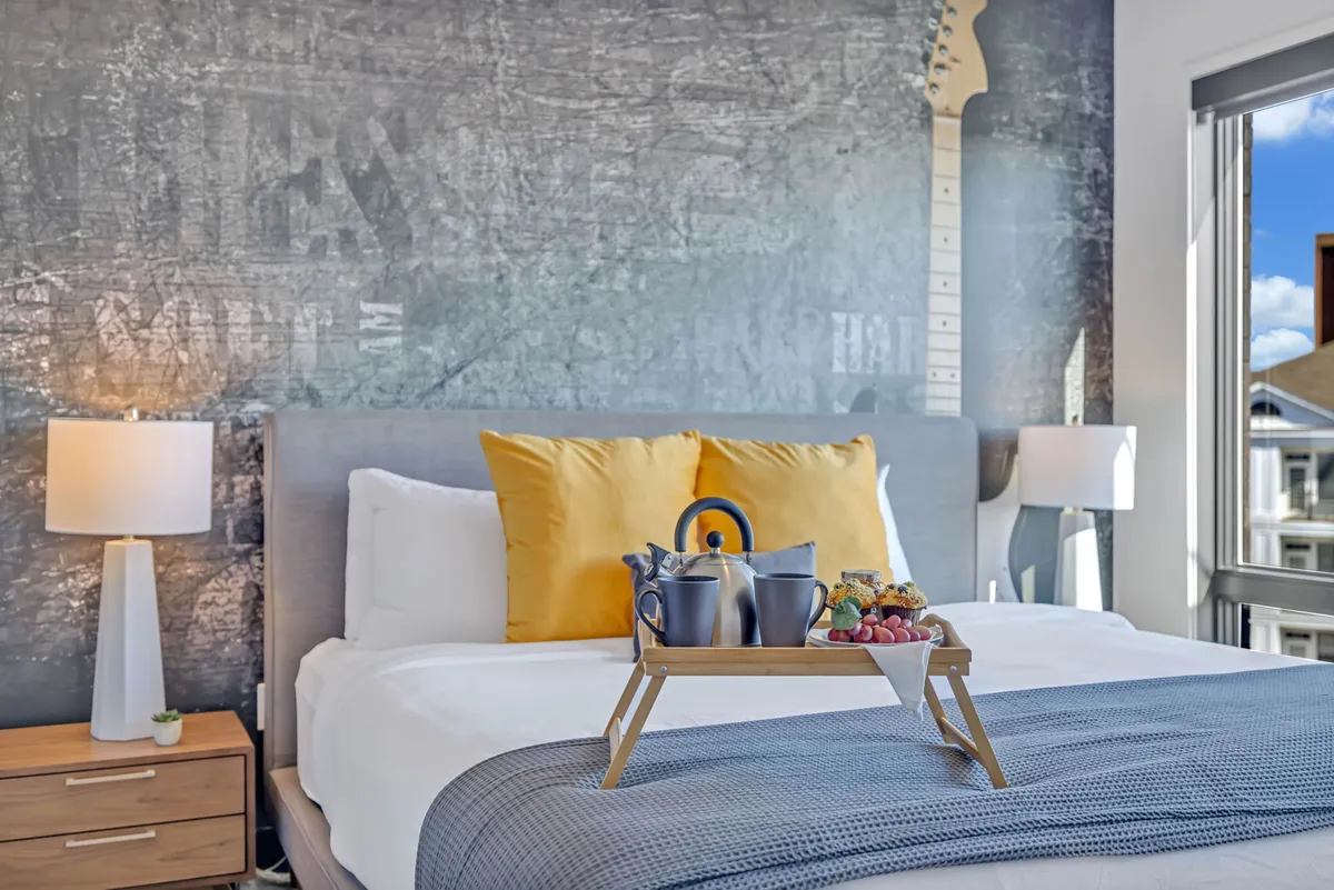 A chic bedroom featuring a bed with yellow accent pillows, a bold guitar mural on the wall, and a breakfast tray atop the bedding. The room feels comforting, stylish, and designed for morning relaxation.