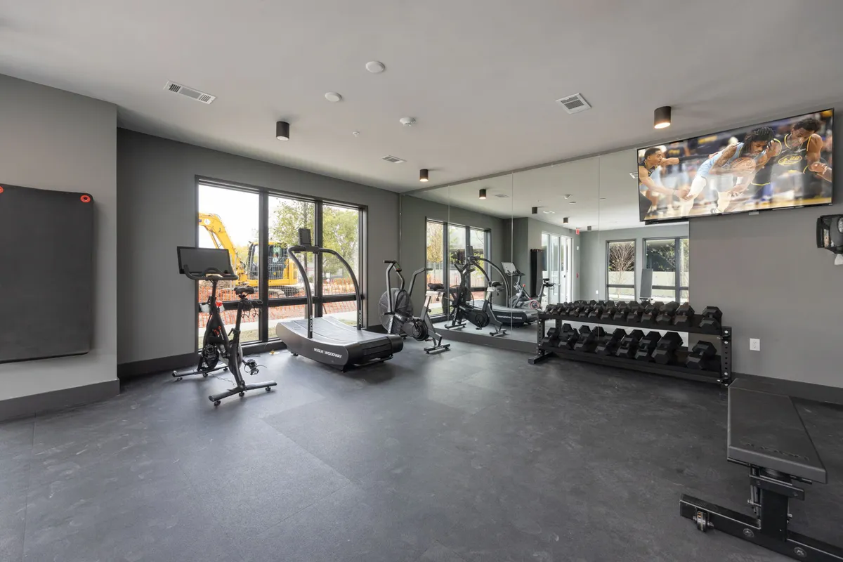 A contemporary fitness center with large windows letting in natural light. Exercise equipment including treadmills, a weight area, and a television screen create an energizing space for both individual and group workouts.